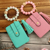A pink and mint silicone PU leather key chain wristlet wallet with beaded bracelets on a wooden surface.