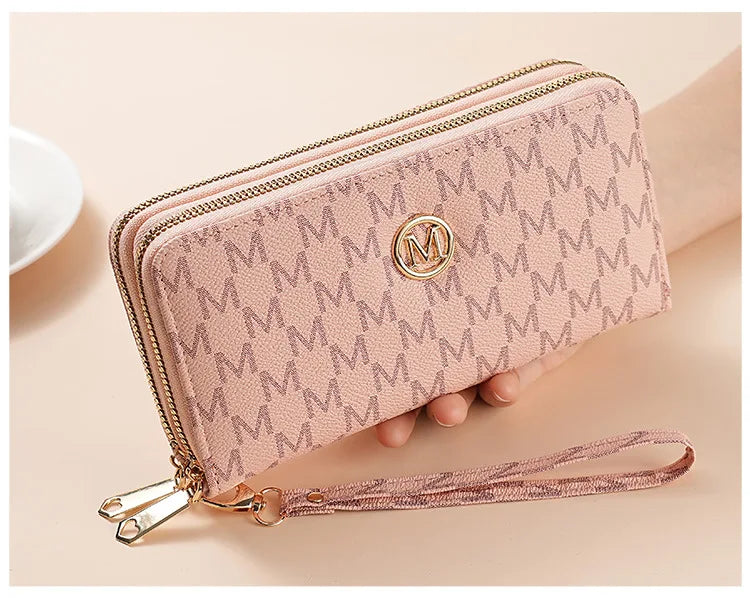 A light pink double zipper wallet with a gold logo and a matching pink strap is shown being held by a hand.
