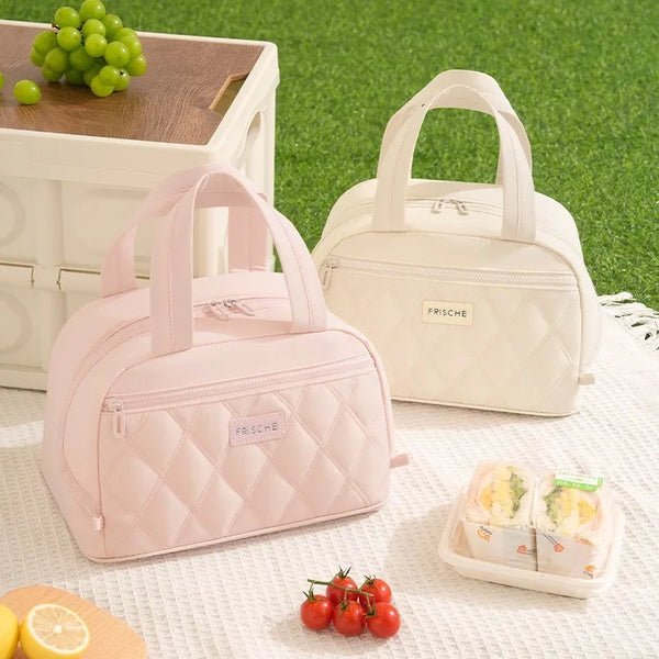 A pink quilted insulated lunch tote bag next to a cream-colored version and some fruit on a white cloth.