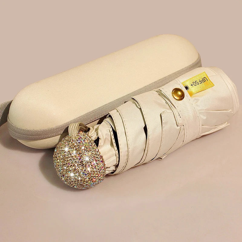 A compact white rhinestone umbrella with a gold-colored UV protection tag is shown lying on a light surface.