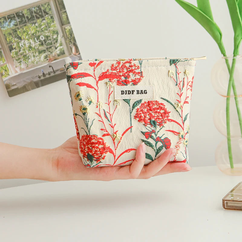 A hand holds a floral canvas makeup bag featuring red and green flowers with the text DIDF BAG printed on it.