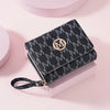 A black letter zipper wristlet wallet with a gold monogram and leather strap resting on a pink surface.