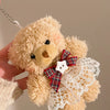 A fluffy beige teddy bear plush toy with a white lace dress and a red plaid bow, being held by a hand.