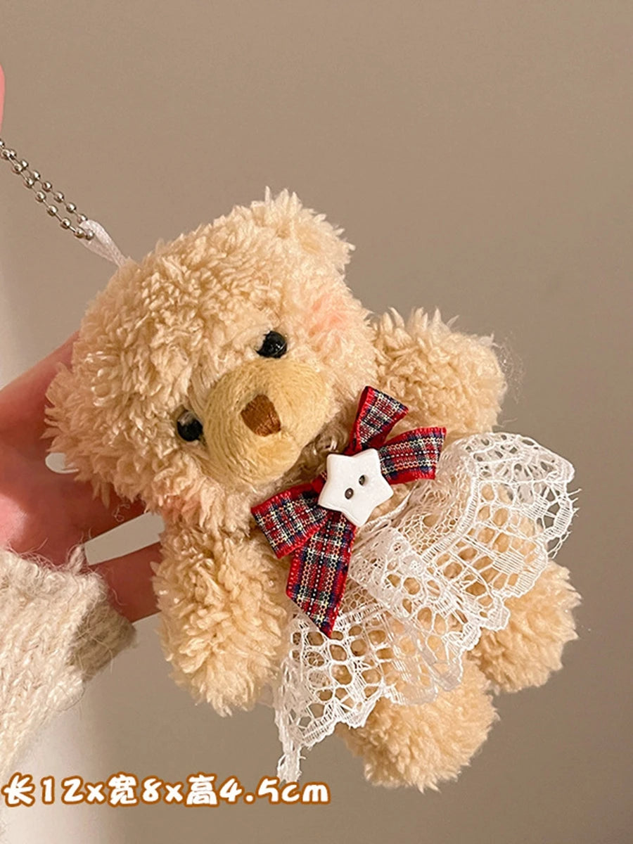 A fluffy beige teddy bear plush toy with a white lace dress and a red plaid bow, being held by a hand.