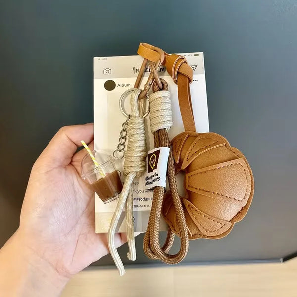 A hand holds a leather croissant keychain lanyard with a pendant and attached bag.