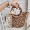 A hand is holding a beige woven rattan beach bag with a lace tied bow against a white background.