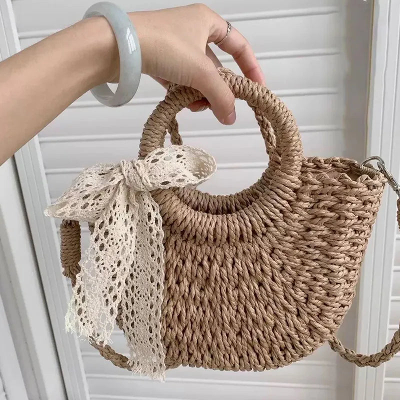 A hand is holding a beige woven rattan beach bag with a lace tied bow against a white background.