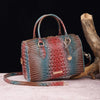 A reddish-brown crocodile pattern leather crossbody tote bag resting on a light surface next to a piece of driftwood.