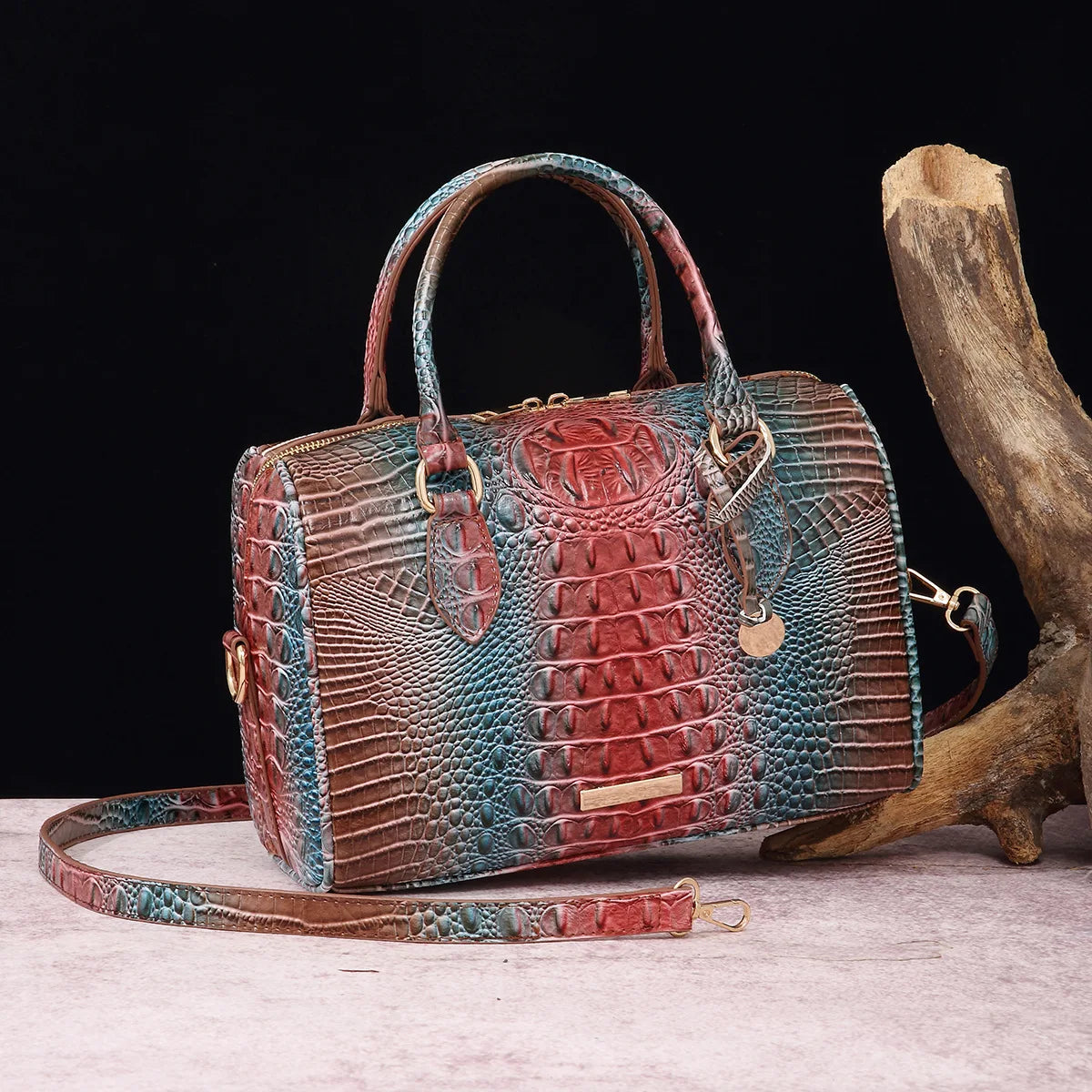 A reddish-brown crocodile pattern leather crossbody tote bag resting on a light surface next to a piece of driftwood.