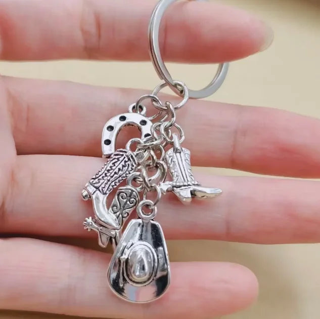 Silver keychain set featuring a cowboy boot, hat, and horseshoe charms held by a hand.