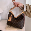 A hand holds a brown leather crossbody tote bag with a gold chain and a small leather tag against a white background.