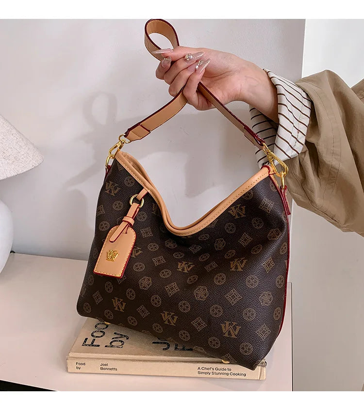 A hand holds a brown leather crossbody tote bag with a gold chain and a small leather tag against a white background.