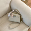 A light beige PU leather crossbody clutch bag with a woven strap resting on a cream upholstered surface.