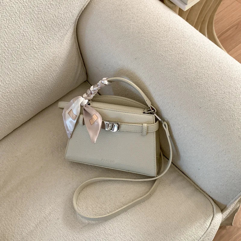 A light beige PU leather crossbody clutch bag with a woven strap resting on a cream upholstered surface.