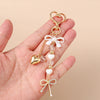 A hand holds a gold-toned pearl bow heart keychain with a loop and dangling elements.