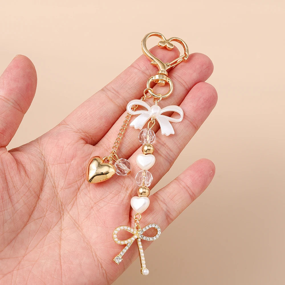 A hand holds a gold-toned pearl bow heart keychain with a loop and dangling elements.