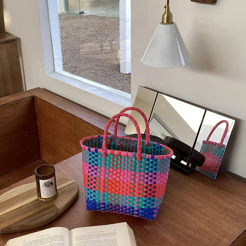 A vibrant hand woven PVC vegetable basket tote bag with pink, blue, and green stripes sits on a wooden table.