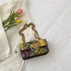 An elegant retro crossbody bag with a patterned fabric and gold chain rests on a white linen cloth next to pink and yellow tu