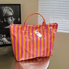 A bright orange and pink striped tote bag is displayed against a window with horizontal blinds.