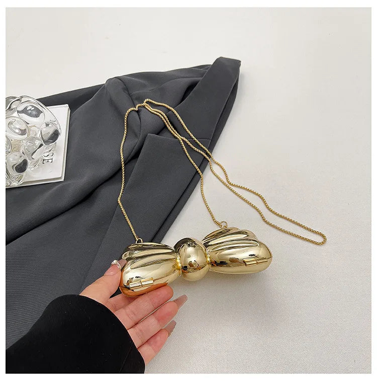 A hand holds a gold-colored acrylic bow clasp mini handbag with a delicate chain strap against a dark gray fabric background.