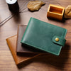 Portefeuille pliable en cuir vert foncé posé sur une surface en bois à côté d'autres articles en cuir et d'une petite boîte e