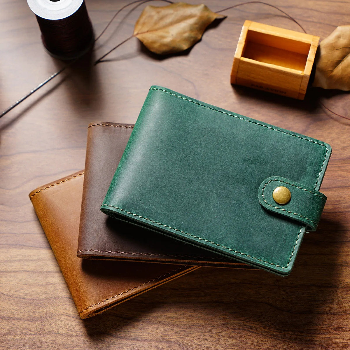 Portefeuille pliable en cuir vert foncé posé sur une surface en bois à côté d'autres articles en cuir et d'une petite boîte e