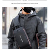 A person wearing a gray hooded jacket is carrying a black anti-theft crossbody chest bag on their chest.