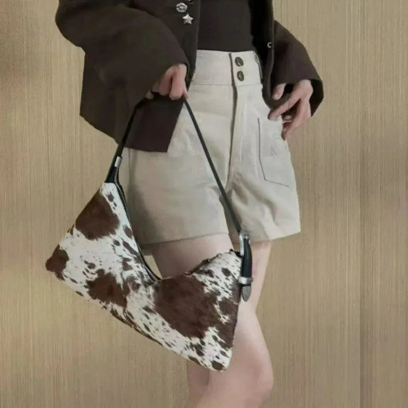 A woman wearing a brown jacket and beige shorts holds a brown and white cowhide crossbody bag.
