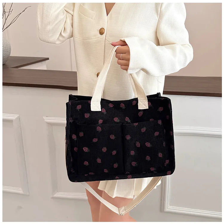 A person holds a black canvas tote bag with a strawberry pattern and light beige handles.
