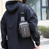 A person wearing a black jacket is carrying a dark gray Oxford sling chest bag with a phone pocket on their shoulder.