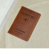 A brown genuine leather passport holder with a stitched edge and embossed text on a beige fabric background.