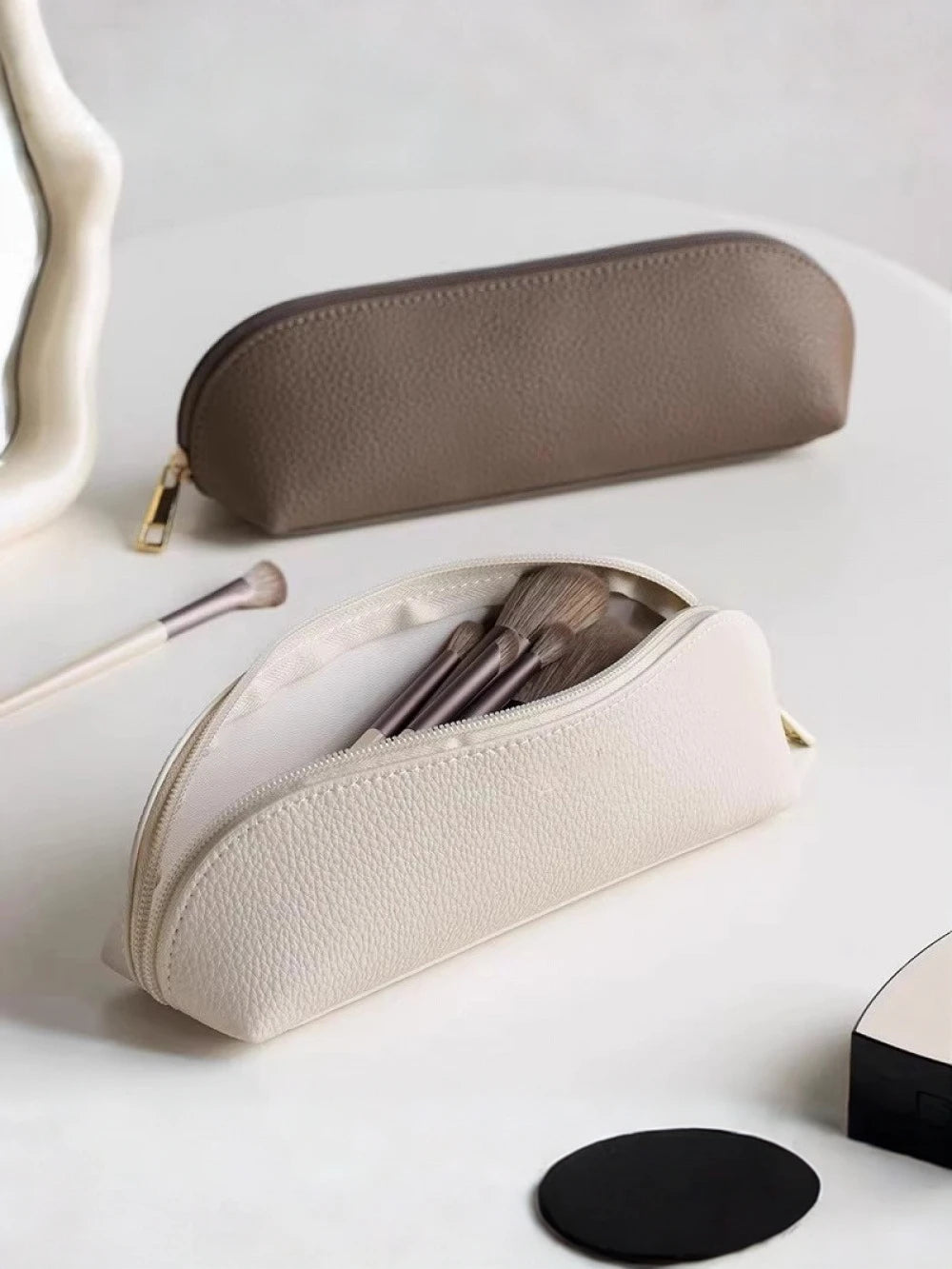 A beige makeup pouch and a gray cosmetic pencil case are displayed on a white surface.