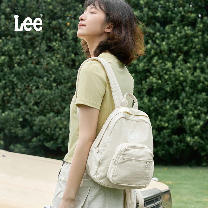 A young woman wearing a light green shirt and overalls carries a beige lightweight waterproof daypack outdoors.