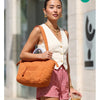 A woman wearing a white top and red plaid shorts holds an orange quilted tote bag.