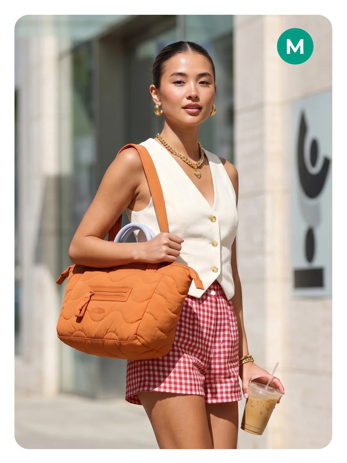A woman wearing a white top and red plaid shorts holds an orange quilted tote bag.
