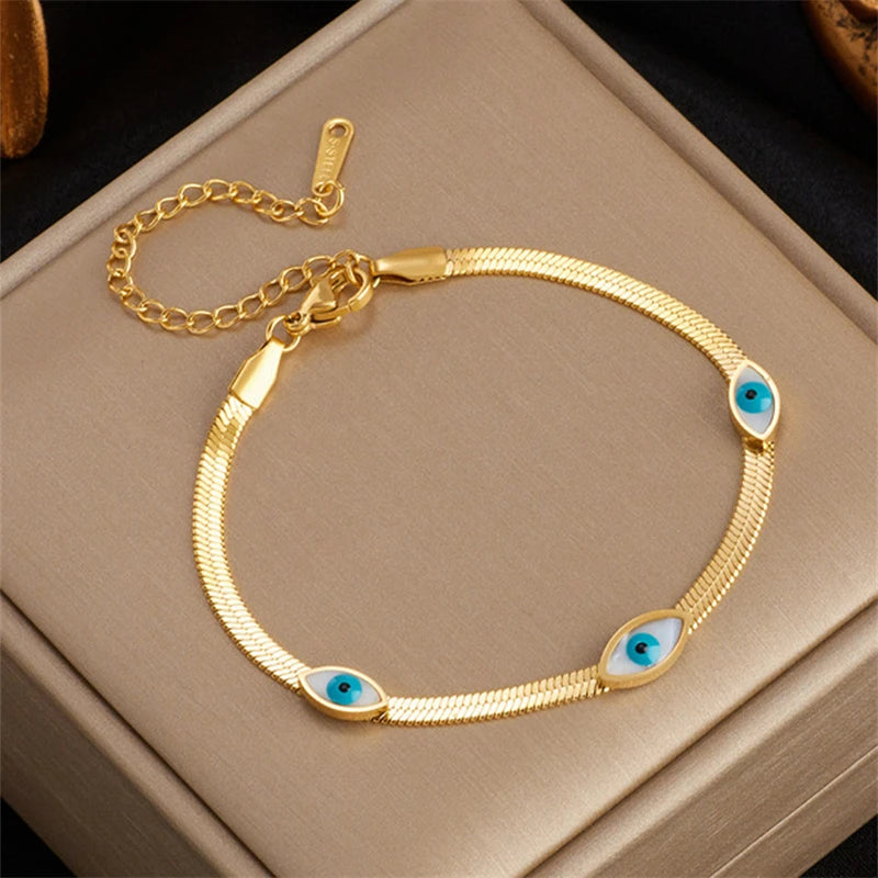 Gold evil eye crystal cuff bracelet with a chain and decorative stones resting on a beige box.