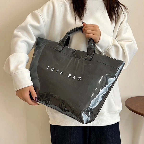 A person holds a black glossy PVC tote bag against a white sweatshirt.