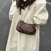 A brown leather shoulder bag with a gold chain and red cherry pendants is carried by a person wearing a light beige coat.