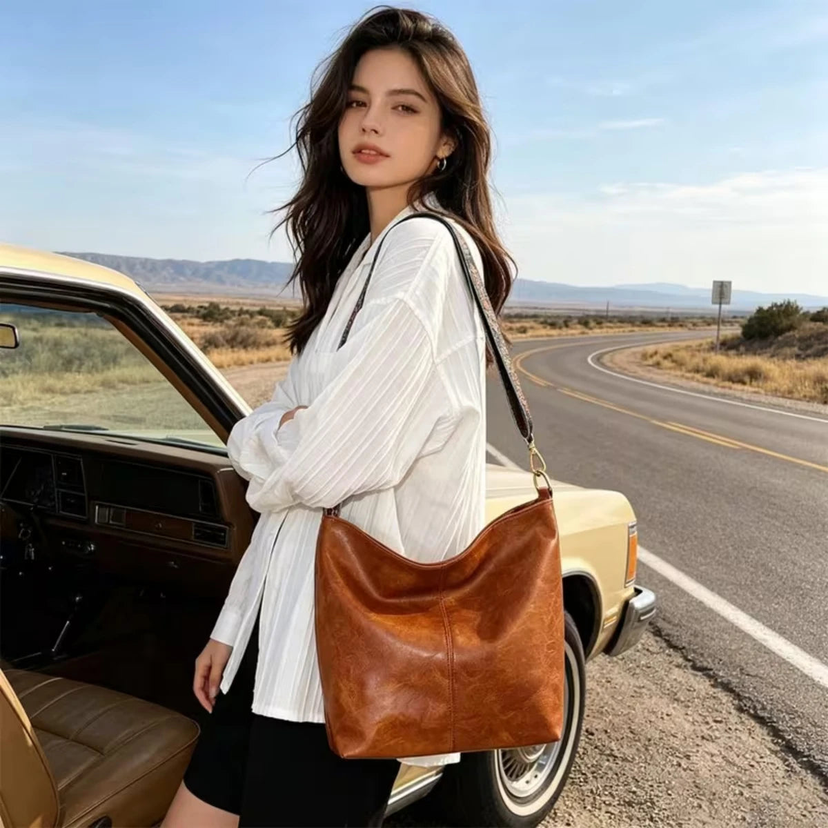 A woman stands next to a gold car with the open door, holding a brown leather crossbody bag.
