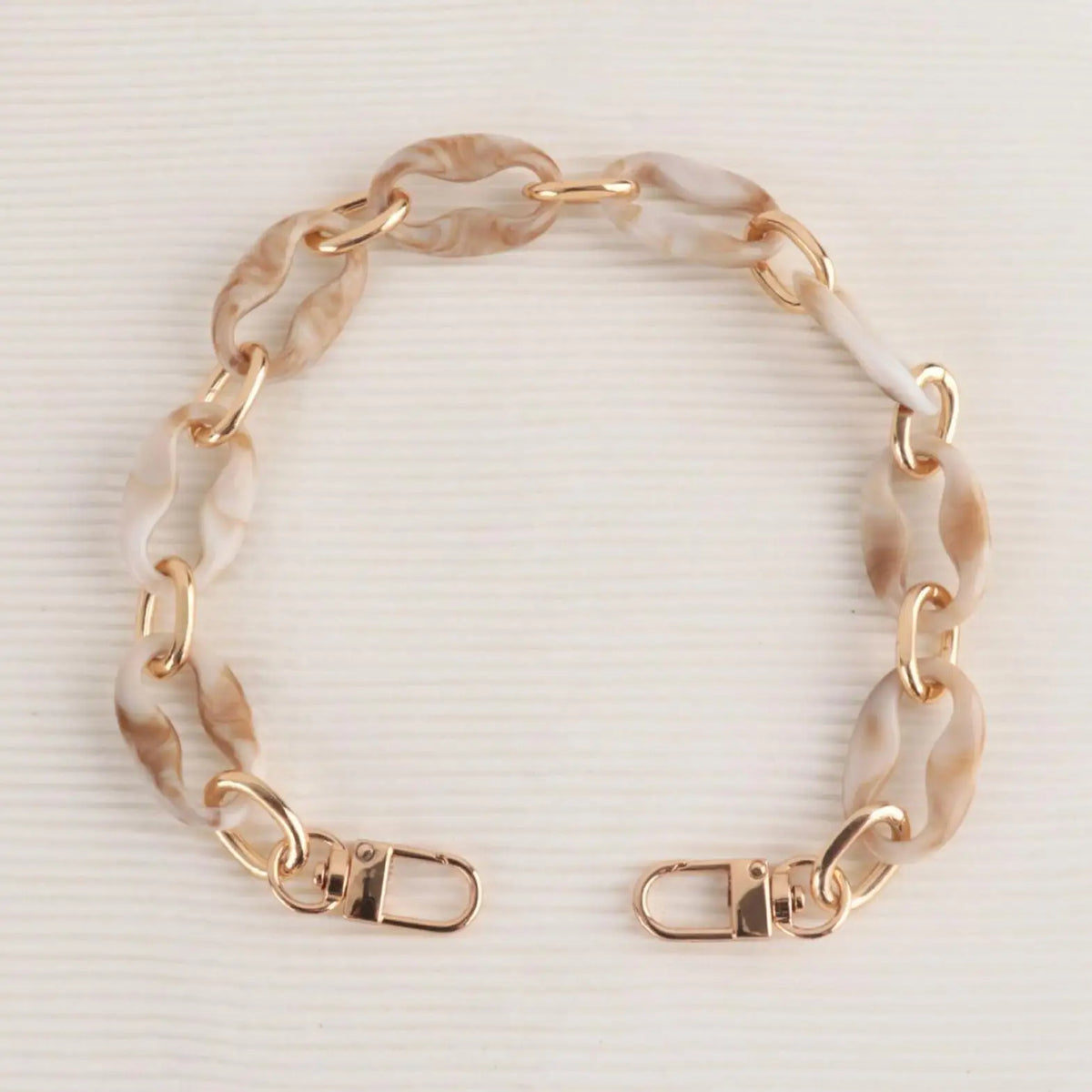 A delicate gold-toned chain strap made of marbled beige and white acrylic is displayed against a white background.