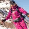 A woman in a pink jacket and black hat is hiking on a snowy mountain slope, carrying a multi-pocket waist bag.