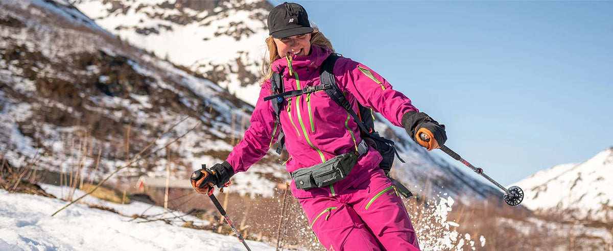 A woman in a pink jacket and black hat is hiking on a snowy mountain slope, carrying a multi-pocket waist bag.