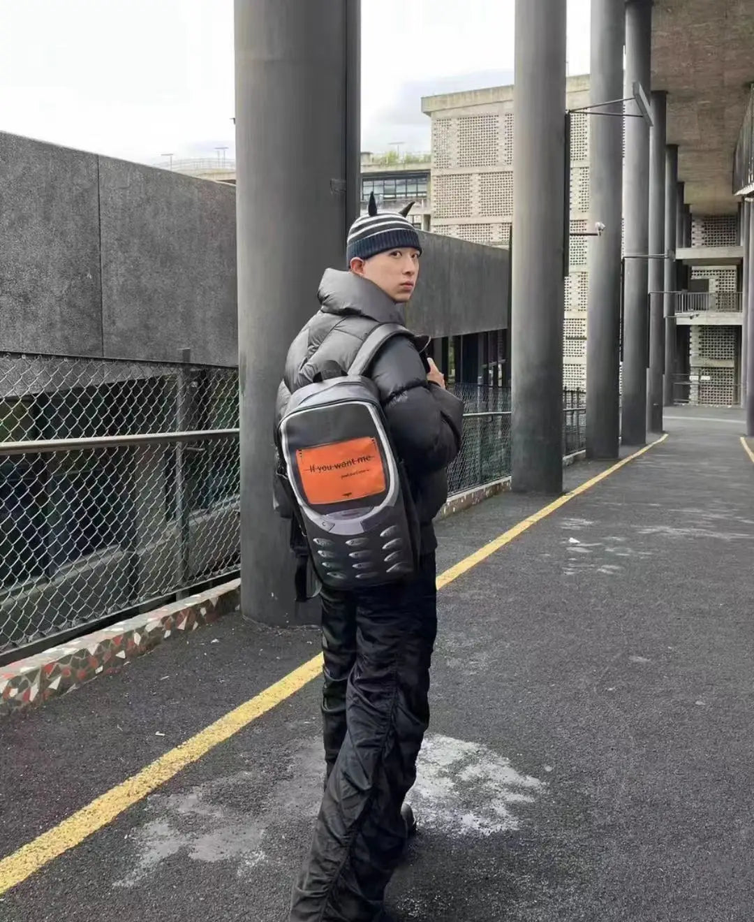 A person wearing a black padded jacket and a vintage Y2K telephone backpack walks down an outdoor walkway.