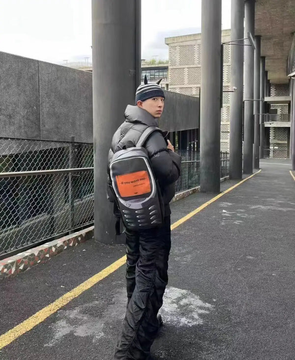 A person wearing a black padded jacket and a vintage Y2K telephone backpack walks down an outdoor walkway.