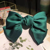 A dark emerald green silk bow hair clip displayed on a white surface with greenery.