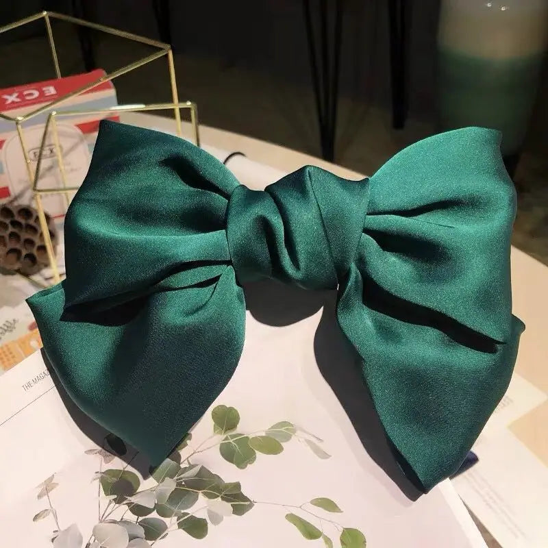 A dark emerald green silk bow hair clip displayed on a white surface with greenery.