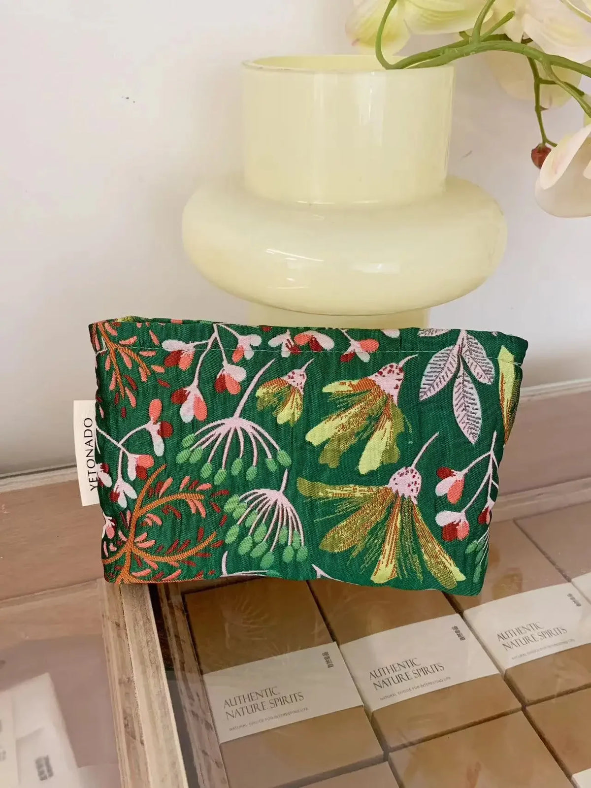 A dark green cosmetic pouch with floral embroidery rests on a light-colored ceramic stand.