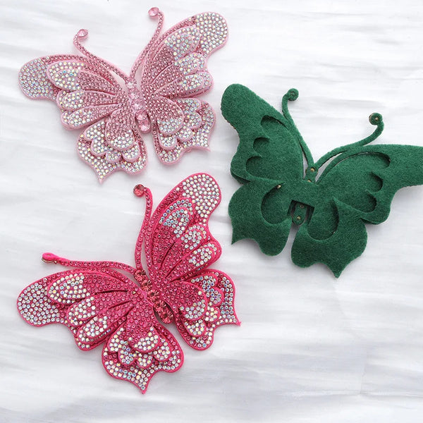 Zwei dekorative Rhinestone-Schmetterlingsapplikationen in rosa und grün auf weißem Stoff.