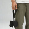 A hand is holding a black nylon zipper wristlet pouch with a small chain attached, next to olive green pants.