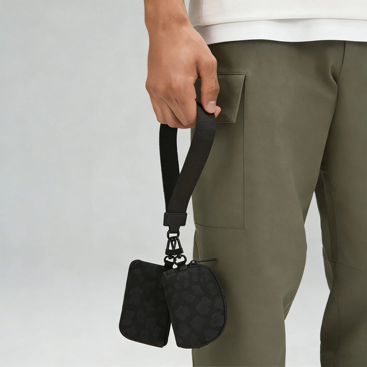 A hand is holding a black nylon zipper wristlet pouch with a small chain attached, next to olive green pants.